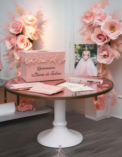 Pink Paris quinceanera theme lluvia de sobres box sitting on top of a table with a picture of the quinceañera in a pink dress, and envelopes with a gold pen on the table. roses and pink pampas decor in the background.