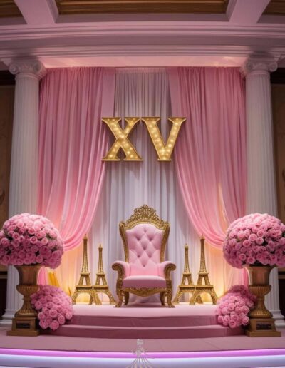 Pink Paris quinceanera theme throne with letter XV at the center above the pink and gold throne. the letters are lit up. There is soft sheer pink drapery to sides and white drapery to the back of the throne. pink bouquets sit on gold vases and the throne sits on top on two steps with golden eiffer towers to each side.