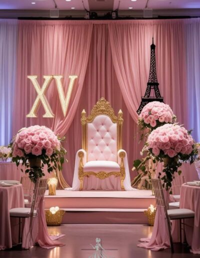 Pink Paris quinceanera theme throne in light pink with the letters VX to the left in gold and a black eiffel tower to the right. pink roses bouquets on both sides of the pink and gold throne and chairs and tables to each side both in light pink and light pink drapery.
