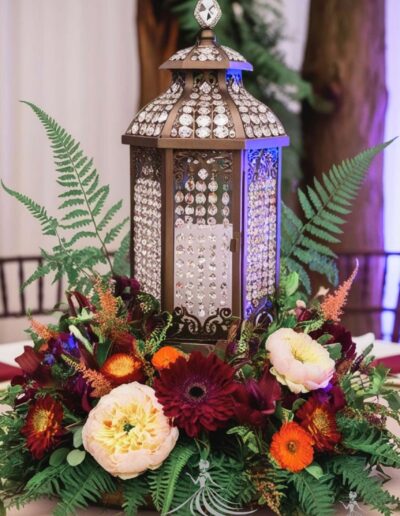 Enchanted forest centerpiece lantern dark theme features darker tones of flowers such as burgundy, deep orange, and greens. the lantern is a dark brassy color decorated by clear crystal beads.